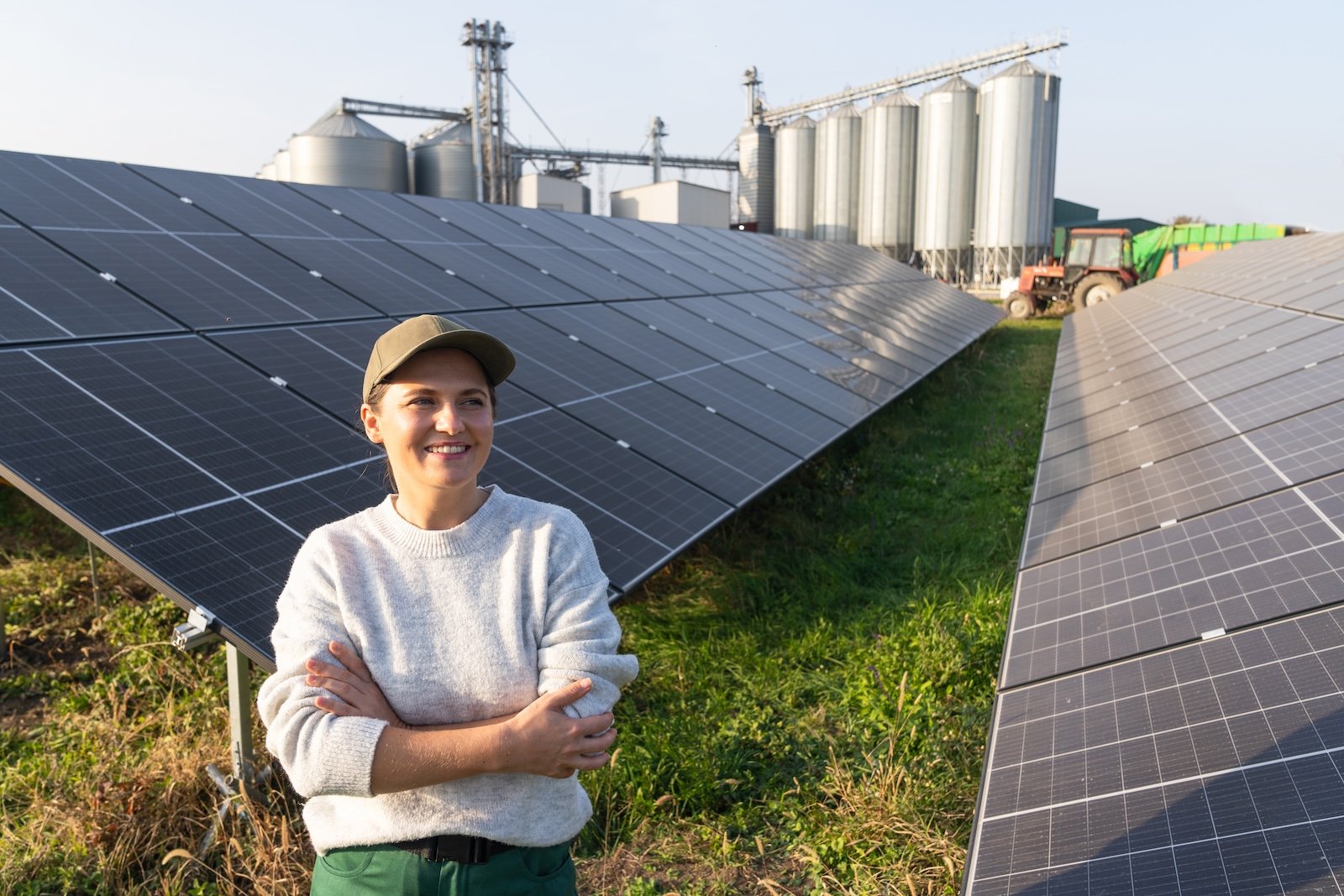 Lachende boerin bij een grondopstelling zonnepanelen, met graansilo's en tractor op de achtergrond